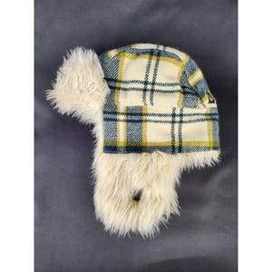 May include: A winter hat with ear flaps. The hat features a plaid pattern in cream, yellow, and navy blue. The ear flaps and the inside of the hat are lined with a soft, cream-colored faux fur. A small logo is visible on the side.