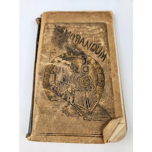 May include: Vintage brown leather-bound book with the title "THE MORANDUM" and an illustration of a steam train. The cover is aged and worn, with visible creases and wear. The book has a black spine and a small, torn corner.