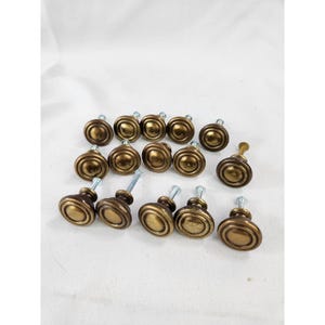 May include: A collection of thirteen round cabinet knobs with a metallic antique brass finish. Each knob features a concentric circular design with a raised center and a flat metal screw extending from the back for mounting purposes.