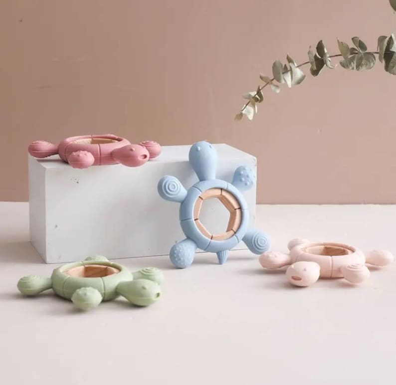 May include: Four colorful turtle-shaped baby teethers. The teethers are in shades of pink, blue, and green, with a wooden ring in the center. The teethers are designed with textured surfaces for soothing gums.