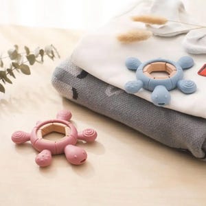 May include: Two turtle-shaped baby teethers. One teether is pink, the other is blue. Both have a wooden ring in the center and soft, textured silicone bodies. The teethers are on a light-colored surface with folded baby clothes in the background.