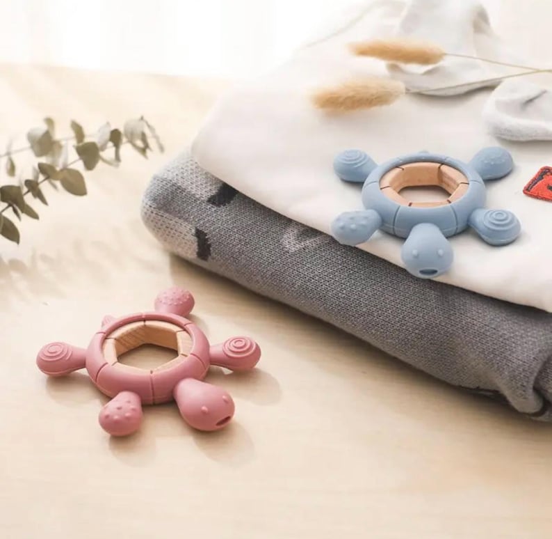 May include: Two turtle-shaped baby teethers. One teether is pink, the other is blue. Both have a wooden ring in the center and soft, textured silicone bodies. The teethers are on a light-colored surface with folded baby clothes in the background.