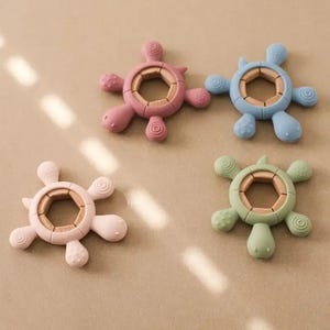 May include: Four colorful turtle-shaped baby teethers. Each teether has a wooden center and a soft, textured silicone exterior in pink, blue, green, and light pink. The turtle's head and flippers are rounded.