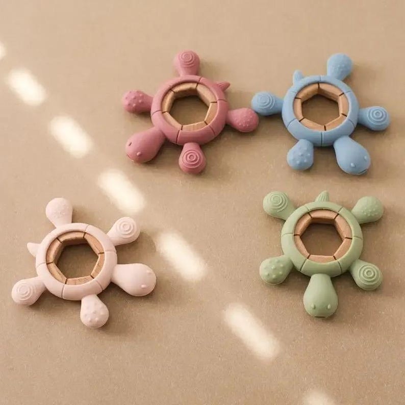 May include: Four colorful turtle-shaped baby teethers. Each teether has a wooden center and a soft, textured silicone exterior in pink, blue, green, and light pink. The turtle's head and flippers are rounded.