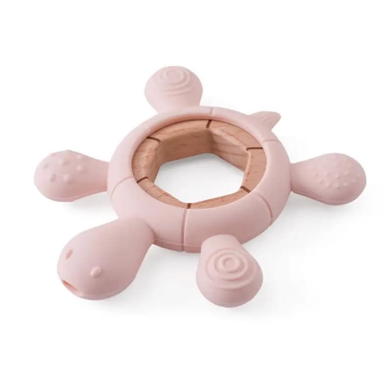 May include: A light pink baby teether shaped like a turtle. The teether has a wooden center and soft, textured silicone flippers and head. The design is intended for babies to grasp and chew on. The teether is designed to soothe sore gums.