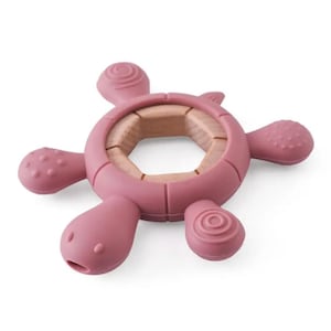 May include: A pink turtle-shaped baby teether with a wooden center. The teether has four textured flippers and a head with a small opening. The center is a light brown color. The teether is made of soft, flexible material.