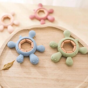 May include: A collection of colorful baby teethers shaped like turtles. The teethers are in shades of blue, green, and pink, with a wooden-colored center. The turtle-shaped teethers are arranged on a wooden tray.