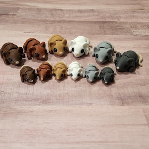 May include: A collection of handmade polymer clay mice in various colors, including brown, tan, white, gray, and black. The mice are arranged on a light brown wooden surface. Each mouse has small, detailed features.