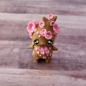May include: A handcrafted brown clay figurine of a creature with large eyes and pink flower accents. The figurine has a stylized design with a floral crown and pink flower details on its body. The background is a light brown wooden surface.