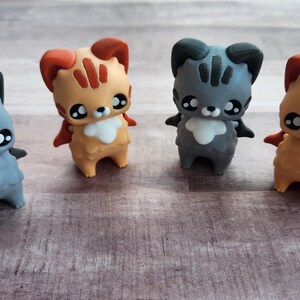 May include: Four small, stylized figurines of cat-like creatures. Two are gray with black accents, and two are orange with red accents. Each has large eyes, small wings, and a fluffy chest. They stand on a light brown wooden surface.