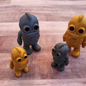 May include: Four small robot figurines in shades of gold and gray are arranged on a wooden surface. The robots have large, round eyes and a simple, blocky design. The lighting highlights the textures and shapes of the toys.