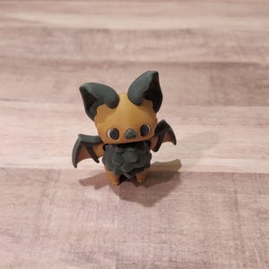 May include: A small, brown and black bat figurine with large, round eyes. The bat has wings that are spread out and a small, furry body.