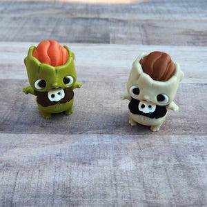 May include: Two small, whimsical figurines. One is green with an orange top and a brown chest with a white skull. The other is off-white with a brown top and a black chest with a white skull. Both have large eyes.