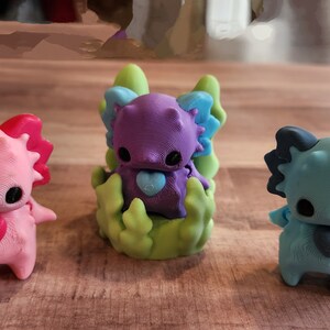 May include: Three small, colorful figurines of axolotls. The axolotl on the left is pink with a red heart, the middle axolotl is purple with a blue heart, and the axolotl on the right is blue with a dark blue heart. The middle axolotl is sitting on a green base.
