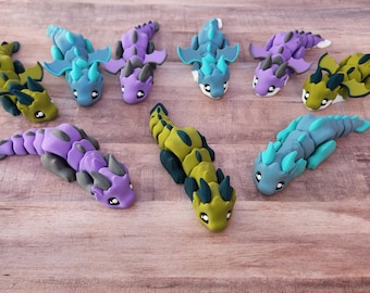 May include: A collection of handmade polymer clay dragon figurines in various colors, including green, blue, and purple. Each dragon has detailed features and is arranged on a wooden surface.