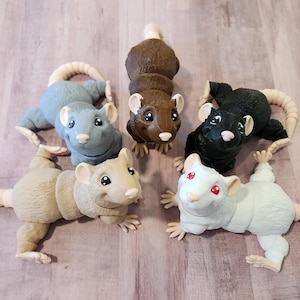 May include: A collection of six rat figurines in various colors: brown, black, white, and shades of gray. Each rat has a long, pale tail and small, detailed paws. The figurines are arranged on a light-colored wooden surface.