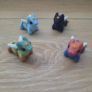 May include: Four small dragon figurines in light blue, dark blue, green-to-orange gradient, and pink-to-blue gradient colors. Each creature features white wings, black eyes, and a small yellow star detail on the forehead, crafted in a smooth finish.