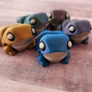 May include: Five small frog figurines in various colors, including blue, purple, gold, and green. The frogs have white eyes and are made of a smooth, shiny material.