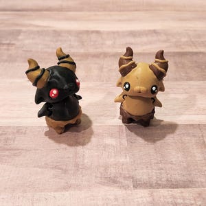 May include: Two small, hand-painted figurines of horned creatures. One is black with red eyes and the other is brown with black horns. Both have a whimsical, cartoonish style.
