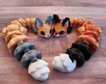 Puede incluir: Una composición en forma de corazón con figuras de zorro estilizadas y elementos en forma de hongo. Los zorros tienen pelaje naranja y negro o azul y naranja. Las setas son de color beige, gris, negro y blanco.