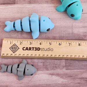 May include: Four articulated dolphin toys, two blue and two grey, are arranged on a wooden surface with a ruler measuring 12 inches. The ruler is labelled "CART3Dstudio".