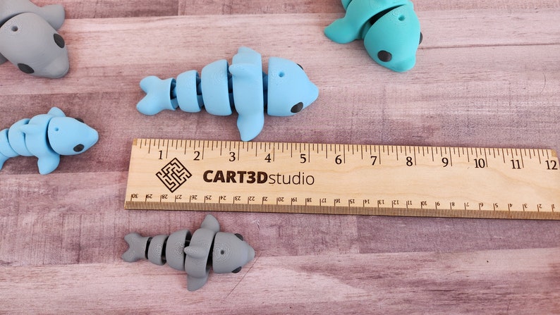 May include: Four articulated dolphin toys, two blue and two grey, are arranged on a wooden surface with a ruler measuring 12 inches. The ruler is labelled "CART3Dstudio".