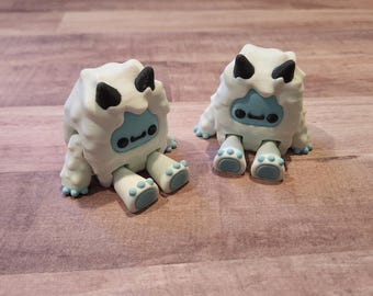 May include: Two white plush toys shaped like snowmen with black eyes and blue mouths. The snowmen are sitting on a wooden surface.