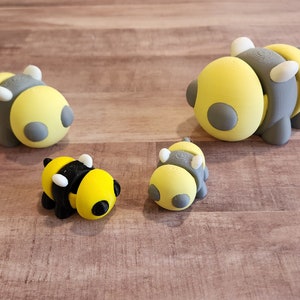 May include: Four small bee figurines made of yellow, gray, and black clay. The figurines are arranged on a wooden surface.