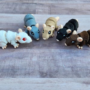 May include: A collection of six miniature rat figurines in various colors, including white, blue, tan, black, and brown. Each rat has a segmented body, a long tail, and small ears. The figurines are arranged on a wooden surface.