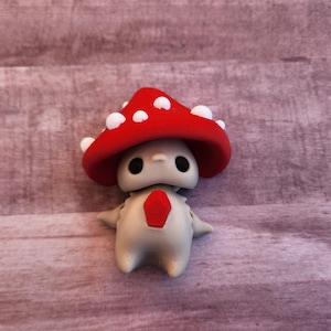 May include: A whimsical figurine of a mushroom-like creature. It has a red cap with white spots, a cream-colored body, and a red diamond-shaped detail on its chest. The figure has large black eyes and small arms.