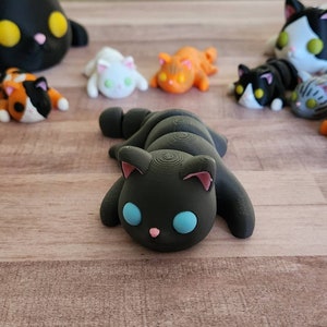 May include: A collection of nine black, white, orange, and gray cat figurines. The figurines are made of plastic and have a cartoonish style. The cats are in various poses, including sitting, lying down, and curled up.