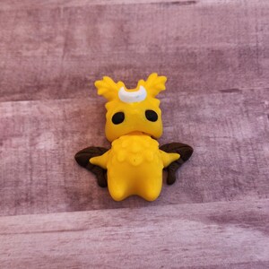 May include: A yellow figurine of a creature with a crescent moon on its head, black eyes, and brown wings. The figurine has a textured body and is set against a wooden background.