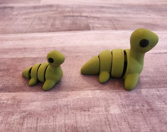 May include: Two green, segmented dinosaur-like figurines with black eyes sit on a wooden surface. One is smaller and in a resting position, while the other is larger and upright. The figures have a smooth, matte finish.