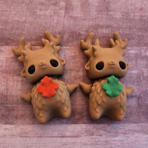 May include: Two whimsical, tan-colored figurines with large black eyes, antlers, and small arms. One has an orange leaf accent, the other a green leaf. The figures are on a textured, purple-brown surface.