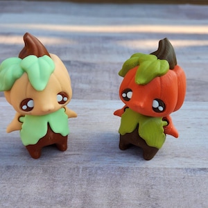 May include: Two small, whimsical pumpkin-themed figurines. One is orange, the other is tan, both with green leaf accents, brown stems, and adorable, wide-eyed faces. They have small arms and legs, giving them a cute, cartoonish appearance.