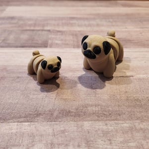 May include: Two small, beige pug figurines. The larger pug is sitting upright with its head tilted slightly to the side. The smaller pug is lying down with its head tilted up. Both pugs have black eyes and noses.