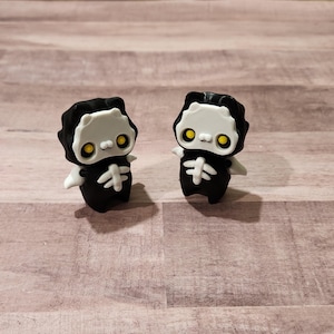 May include: Two small black and white skeleton figurines with yellow eyes. The figurines are standing on a wooden surface.