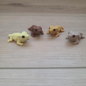 May include: Four small articulated lizard figurines in different colors. One is pale yellow, one is dark brown, one is golden yellow, and one is light gray. Each lizard has a textured body, a wide head, and small black eyes, arranged in a row.