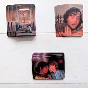 May include: Three stacks of rectangular cards featuring artistic illustrations. One stack shows a person kneeling before a church altar with a crucifix. Another stack depicts a person with dark hair and fangs. The third stack shows two people facing each other.
