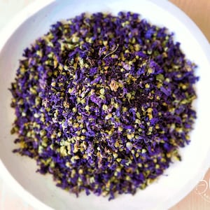 May include: A white ceramic bowl filled with dried purple and green flower buds. The bowl is on a light wood surface.