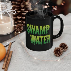 May include: Black coffee mug with a green and yellow dripping text design that says "Swamp Water".