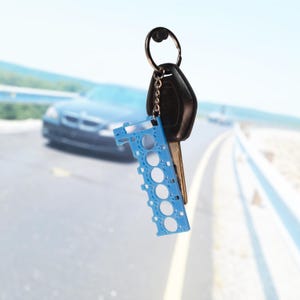 以下が含まれることがあります： 黒いキーフォブとシルバーのキーリングが付いた車のキー。青いエンジン型のキーホルダーに取り付けられています。キーホルダーには一連の円形の切り抜きがあります。背景には車がぼやけています。