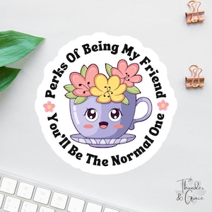 Pode incluir: Adesivo redondo com uma chávena de chá de desenho animado sorrindo, flores e o texto "Perks Of Being My Friend, You'll Be The Normal One". A chávena é roxa, as flores rosa e amarelas.