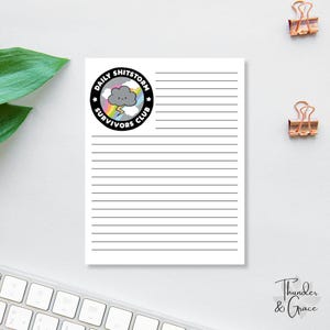 以下が含まれることがあります： 水平な黒い線が入った白いメモ帳と、「Daily Shitstorm Survivors Club」と書かれた円形のグラフィック。グラフィックは雲、虹、稲妻が特徴です。2つの金色のペーパークリップが見えます。