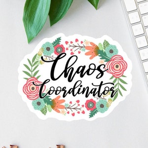 May include: A white sticker with a floral wreath design. The wreath is made up of pink, red, green, and blue flowers. The text "Chaos Coordinator" is written in black cursive lettering inside the wreath.