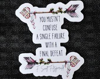 May include: White sticker with two pink and white arrows with floral details. The sticker features the quote "You mustn't confuse a single failure with a final defeat."  - F. Scott Fitzgerald.
