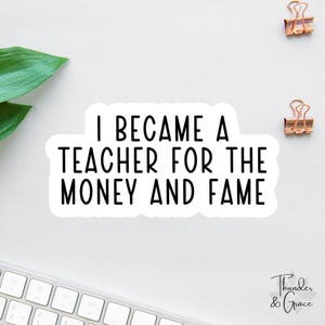 May include: White sticker with the text "I BECAME A TEACHER FOR THE MONEY AND FAME" in black. The sticker is on a white surface with two gold binder clips and a keyboard in the background. A green leaf is in the upper left corner.