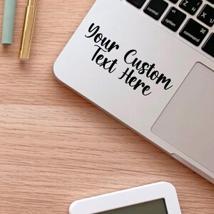 May include: A white laptop computer with a black sticker that says "Your Custom Text Here" on the top of the keyboard. The laptop is on a light brown wooden surface.