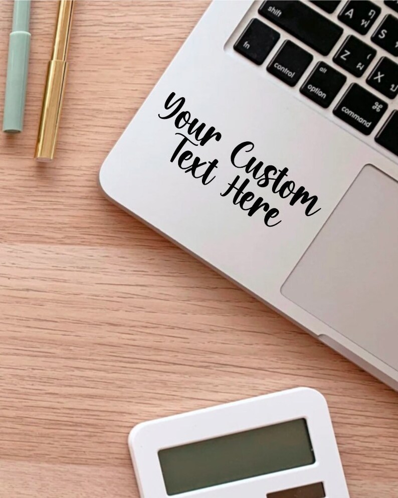 May include: A white laptop computer with a black sticker that says "Your Custom Text Here" on the top of the keyboard. The laptop is on a light brown wooden surface.