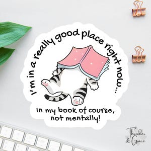 May include: A white sticker with a cartoon cat reading a pink book. The cat's striped legs and paws are visible. Text around the image reads, "I'm in a really good place right now... In my book of course, not mentally!"